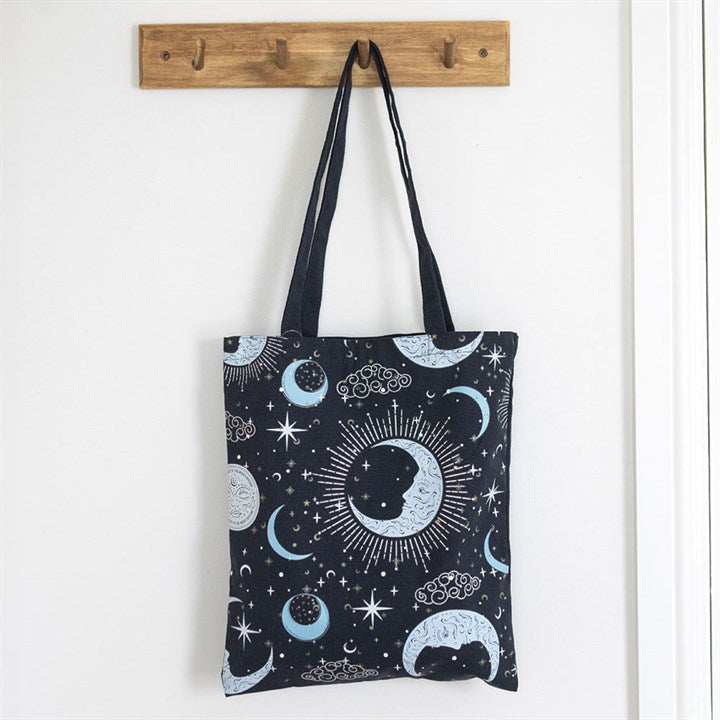 A celestial tote bag featuring pale blue and white moons, stars, clouds, and sunbursts—ideal for carrying your spiritual essentials—hangs from a wooden wall rack against a white wall.