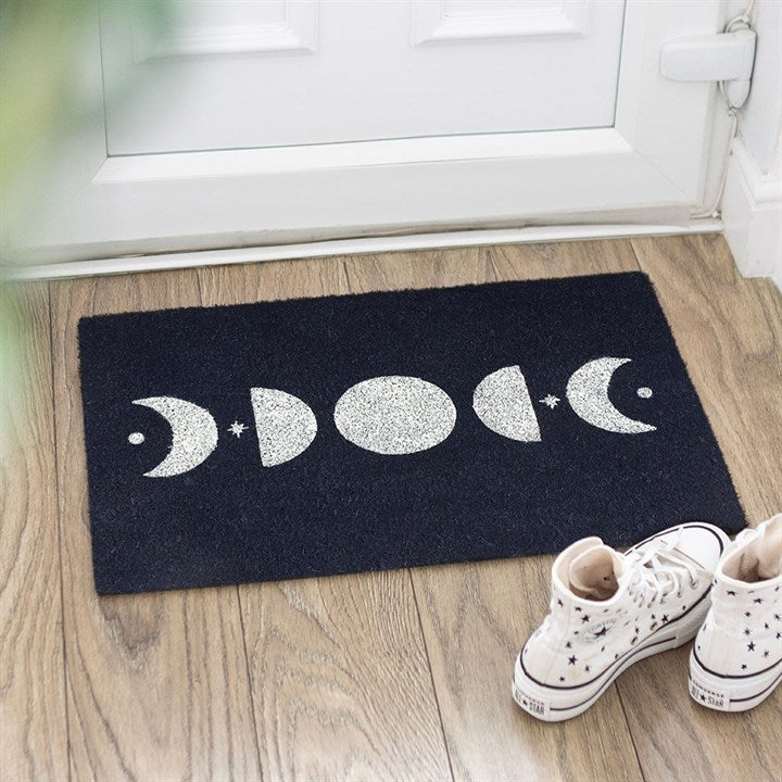 A lunar doormat with white moon phases and stars sits on a wooden floor by a white door, creating a celestial welcome. White high-top trainers with stars and a green plant complete the cosy entrance.