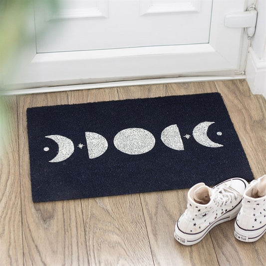 A lunar doormat with white moon phases and stars sits on a wooden floor by a white door, creating a celestial welcome. White high-top trainers with stars and a green plant complete the cosy entrance.