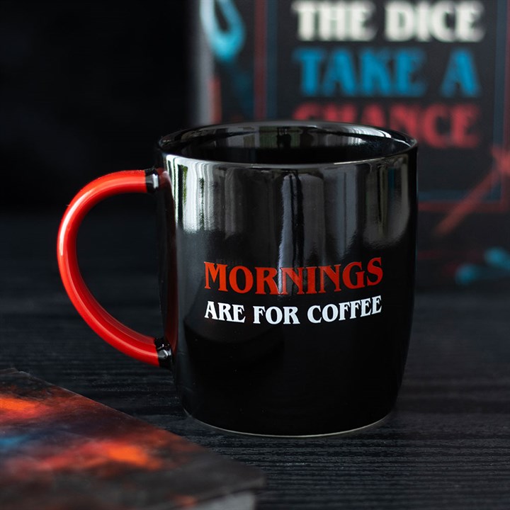A glossy black, microwave-safe mug with a red handle sits on a dark surface. The mug reads “MORNINGS ARE FOR COFFEE” in bold red and white letters. A blurred book cover rests in the background.