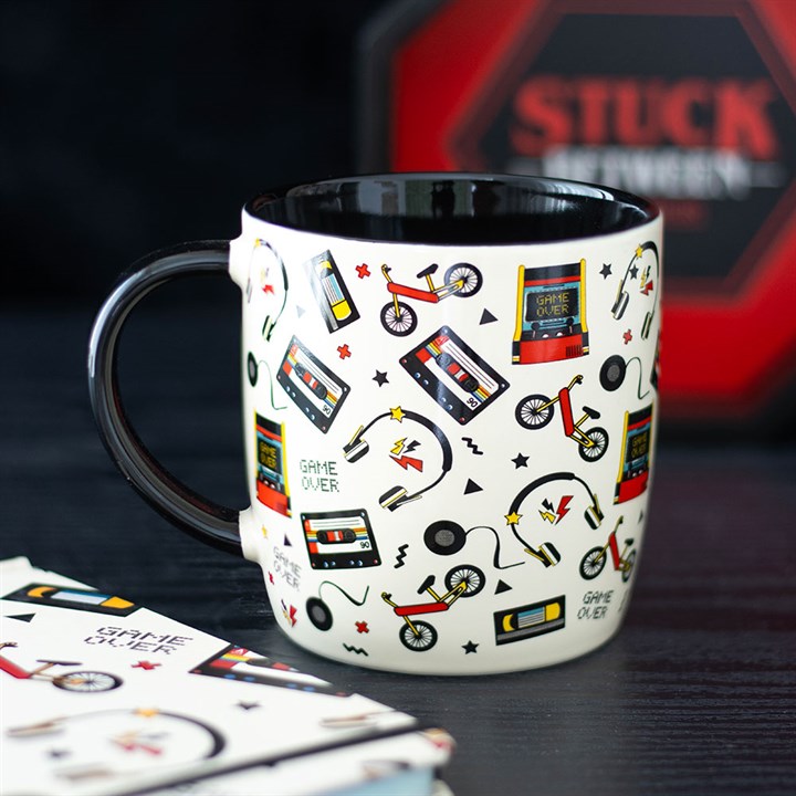 A white classic gaming mug with a black interior features colourful prints of cassette tapes, joysticks, scooters, lightning bolts, and “GAME OVER” messages in a playful, retro style. A blurred red “STUCK” sign can be seen behind.