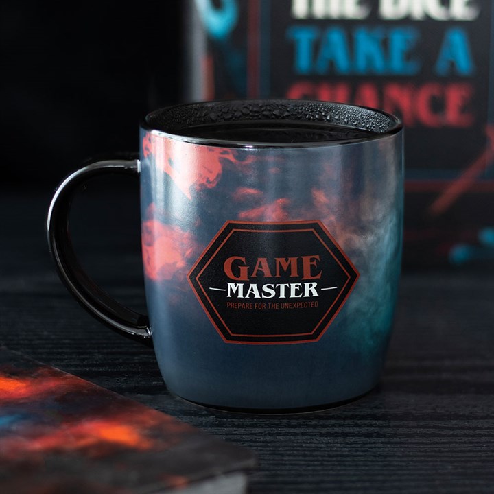 A dark colour-changing mug with red and blue swirling patterns features a hexagonal label reading GAME MASTER, PREPARE FOR THE UNEXPECTED. The gamer mug sits on a dark surface with blurred gaming items in the background.