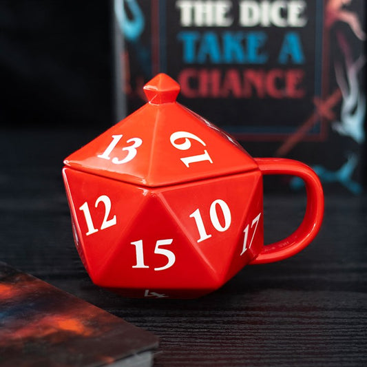 A red D20 mug, shaped like a twenty-sided die, sits on a dark surface; its white numbers and matching lid ready for your next adventure. In the background, a book titled “THE DICE TAKE A CHANCE” is partially visible.