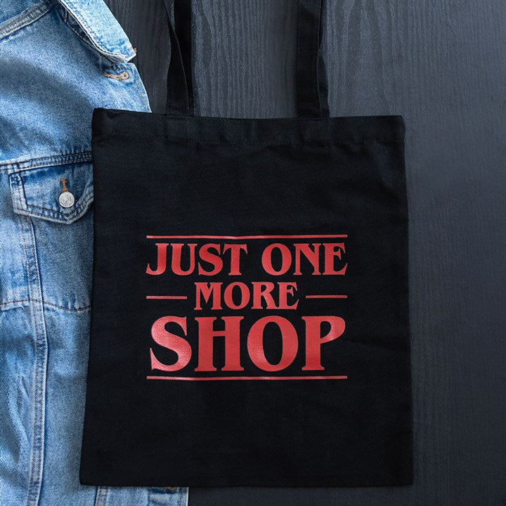 A black tote shopping bag with bold red text reading “JUST ONE MORE SHOP” hangs next to a blue denim jacket against a dark background—perfect for your epic style adventures.