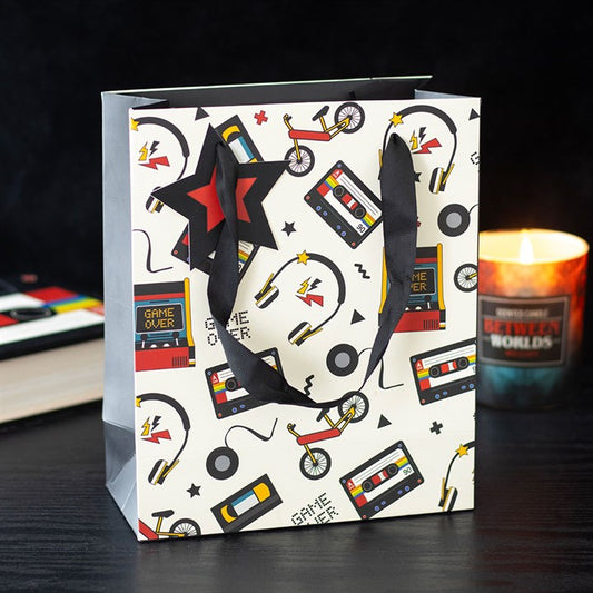 A nostalgic gift bag featuring retro designs such as cassette tapes, bicycles, and headphones, with black ribbon handles and a red-and-black star tag, rests on a dark surface beside a lit candle labelled BETWEEN WORLDS.