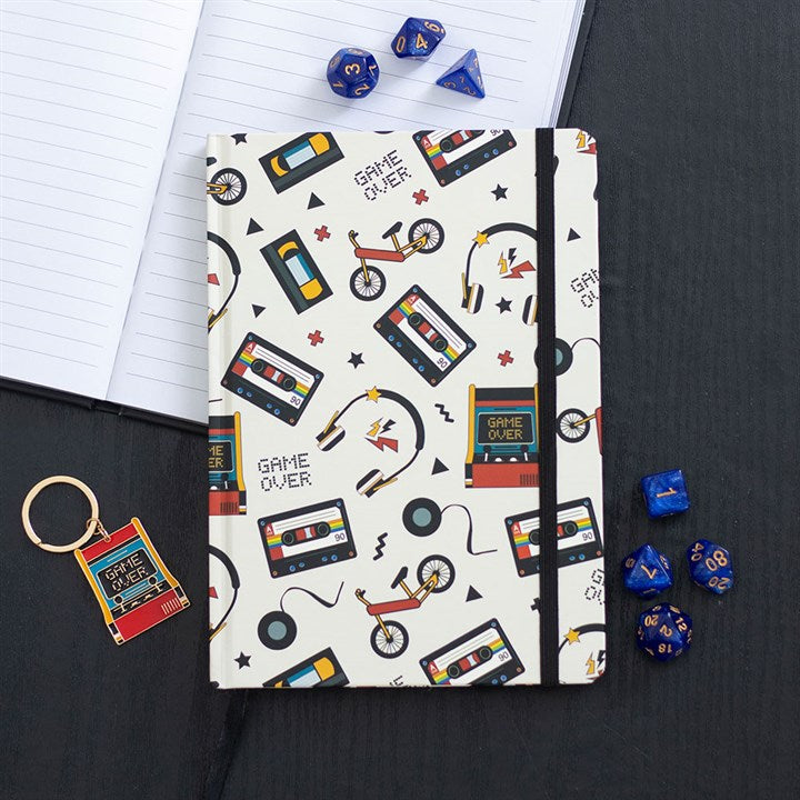 A vintage notebook with a white cover decorated with colourful retro illustrations of cassettes, games consoles, bicycles and headphones sits on a black table beside blue polyhedral dice and nostalgic stationery.