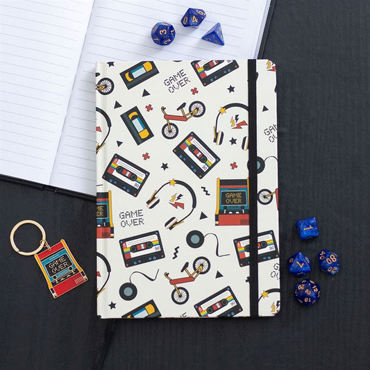 A vintage notebook with a white cover decorated with colourful retro illustrations of cassettes, games consoles, bicycles and headphones sits on a black table beside blue polyhedral dice and nostalgic stationery.
