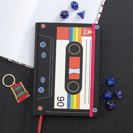 A retro cassette tape notebook with a colourful cover lies on a black table, next to blue polyhedral dice, an open A5 notebook, and a keyring shaped like a red VHS tape labelled Drive Safe—perfect for music lovers.