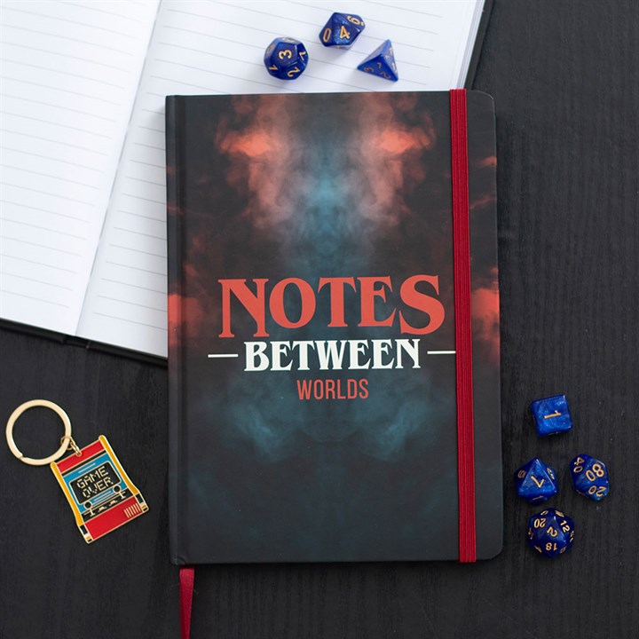 An A5 notebook titled NOTES BETWEEN WORLDS, featuring a smoky design and a red elastic band, lies on a black desk surrounded by blue dice, lined pages in an open notebook, and a vintage Game Boy keyring.