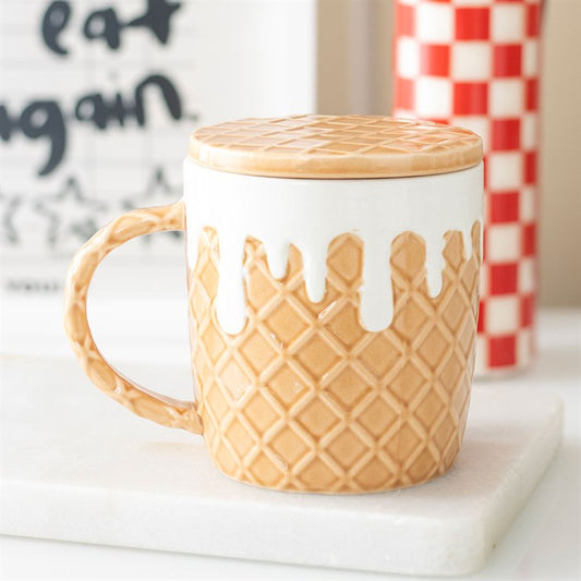 A ceramic coffee mug shaped like an ice cream cone, featuring a mug base with a waffle texture and a lid resembling melting white icing. It rests on a white surface, with a red-checked item and sign in the background.