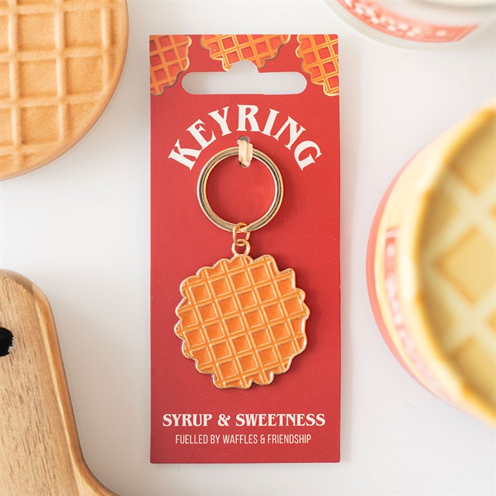 A quirky accessories highlight: a waffle keyring on a red card labelled “Keyring”. Below, it reads “Syrup & Sweetness. Fuelled by Waffles & Friendship.” Perfect as a foodie gift, waffle-themed items surround the card.