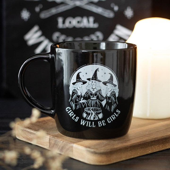 A witch mug showing three witches gathered around a cauldron beneath a crescent moon, with “GIRLS WILL BE GIRLS” below. Ideal as a friendship gift, it rests on a wooden board beside a lit candle.