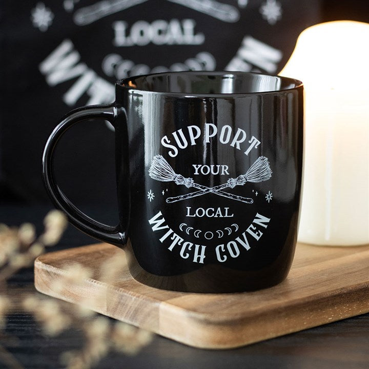 A magical coffee mug with white “Support Your Local Witch Coven” text and crossed broomsticks rests on a wooden tray beside a lit candle, creating a cosy, mystical atmosphere against the softly blurred background.