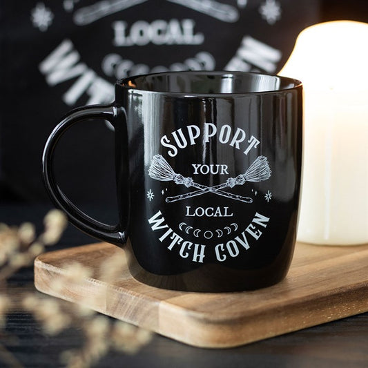A magical coffee mug with white “Support Your Local Witch Coven” text and crossed broomsticks rests on a wooden tray beside a lit candle, creating a cosy, mystical atmosphere against the softly blurred background.