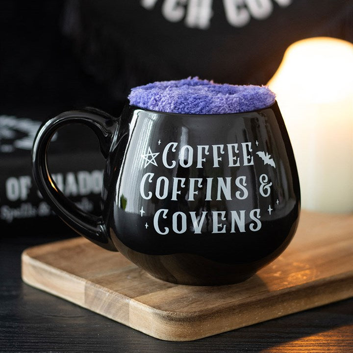 A cauldron-shaped mug with Coffee, Coffins & Covens in white text rests on a wooden board, filled with fluffy purple foam—a perfect witchy gift set, accompanied by a candle and black book glowing gently in the background.