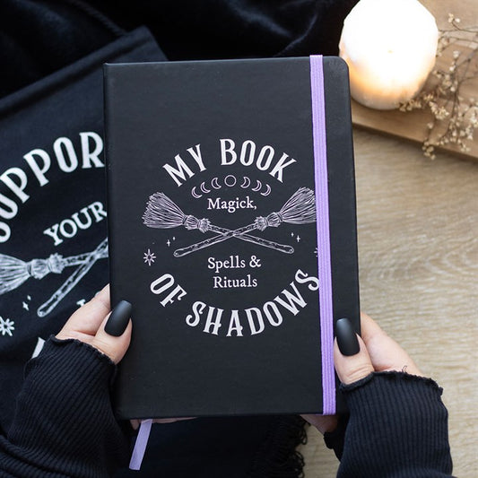 A person with black nail varnish and fingerless gloves holds a witchy notebook titled “My Book of Shadows: Magick, Spells & Rituals,” ideal for jotting down moon ritual notes. A lit candle glows on the wooden surface nearby.
