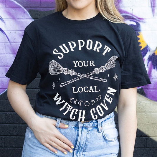 A person wearing a unisex T-shirt bearing the words “Support Your Local Witch Coven”—ideal for alternative wardrobes—stands against a colourful graffiti wall. Only their torso and one hand resting on their hip are visible.