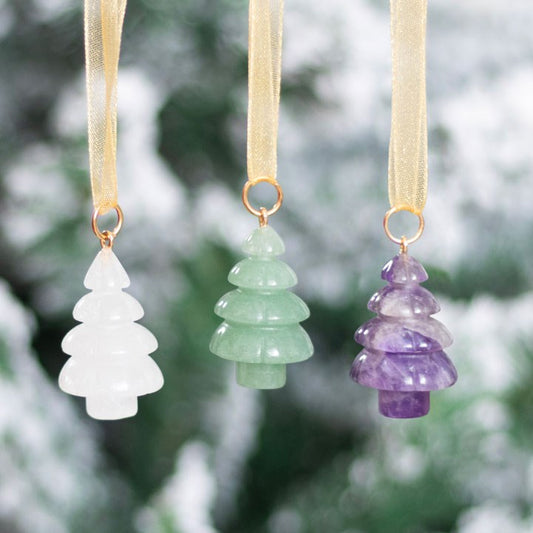 Three crystal Christmas decorations—white, green, and purple—hang from gold ribbons against a blurred, snowy outdoor background with green foliage. Each bauble has a gold ring at the top.
