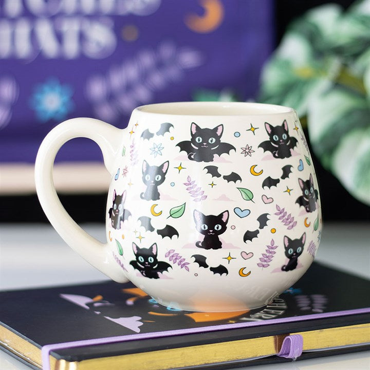 A cream-coloured ceramic mug adorned with charming black cats, bats, stars, moons, hearts, and sprigs of lavender rests on a closed book—perfect for bringing mystical vibes to your kitchen. The blurred background suggests greenery and hints of purple décor.