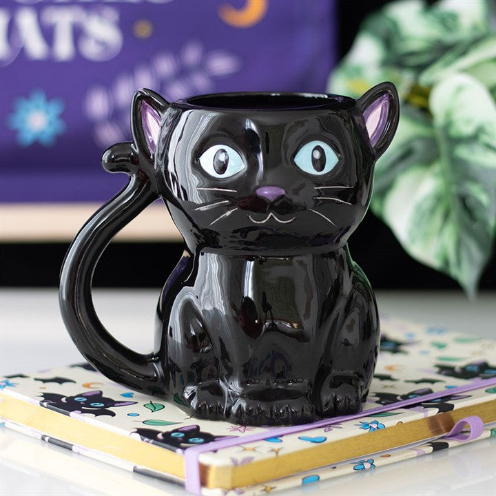 A shiny black cat-shaped ceramic mug with large blue eyes, a curled tail handle, and pink inner ears sits on a notebook decorated with black cat illustrations, adding witchy aesthetics against a blurred leafy plant and purple background.