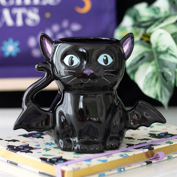 A glossy black cat-shaped ceramic mug with wide blue eyes, a curled tail for a handle, and bat-like wings sits on a cat-patterned notebook—perfect as a spooky gift amidst plants and purple décor in the background.