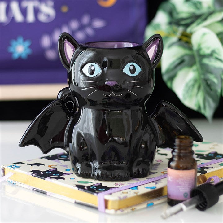 A glossy black bat-shaped cat mug with blue eyes and wings sits on a notebook decorated with black cat faces, adding a touch of gothic décor. A small dropper bottle, leafy plant, and purple starry sign complete the scene.