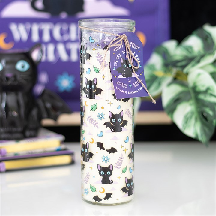 A tall black cat candle adorned with charming cats, bats, moons, and stars. A purple Enchanted Candle tag hangs at the top. In the background are plant leaves, books, and a black cat figurine for gothic home décor.