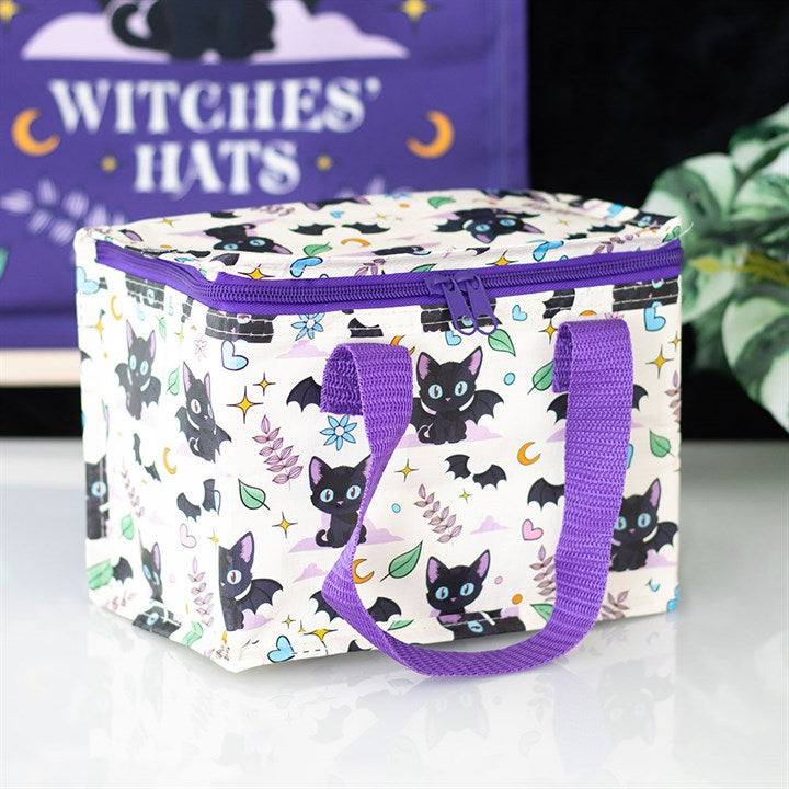 This insulated lunch bag, made from recycled plastic, features a black cat design with cute bats, hearts, moons, and leaves. It has purple handles, a zipped top, and sits on a white surface near a leafy plant.