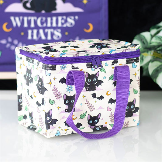 This insulated lunch bag, made from recycled plastic, features a black cat design with cute bats, hearts, moons, and leaves. It has purple handles, a zipped top, and sits on a white surface near a leafy plant.