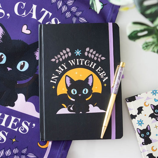 A black A5 notebook featuring a charming black cat illustration with blue eyes and the phrase “In My Witch Era”. The cat is seated beneath purple branches with a crescent moon above. Displayed on a purple cloth beside an amethyst chip pen and another cat-themed notebook.