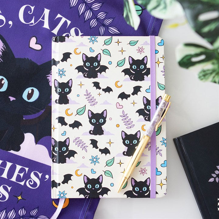 An A5 notebook with a black cat print featuring bat wings, moons, stars, and purple accents lies on the table. An amethyst chip pen rests on top of it, surrounded by cat-themed items and a leafy plant.
