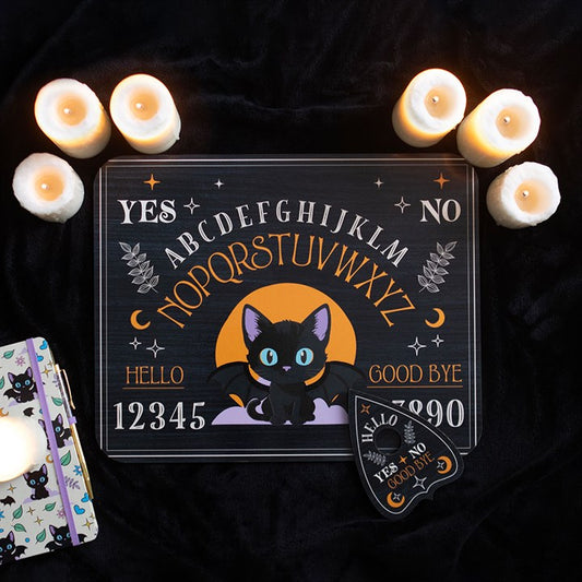 A charming spirit board featuring a cartoon black cat, moon, stars, alphabet, numbers, and the words YES, NO, HELLO, and GOODBYE rests on black fabric, surrounded by six lit candles and a matching planchette.