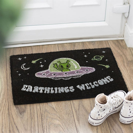 A sci-fi doormat showing a green alien in a purple UFO, with stars and planets. “EARTHLINGS WELCOME” is written on a black background. White trainers sit next to the mat in front of a white door.