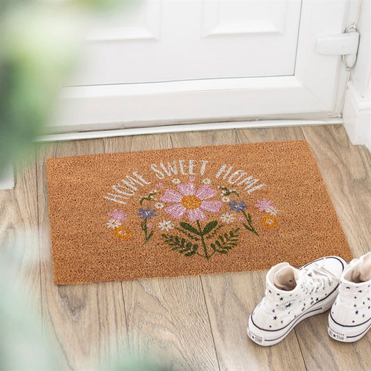 A ‘Home Sweet Home’ doormat with colourful flowers brightens this spring doorstep décor, set on wooden flooring by a white door. White high-top trainers and blurred green leaves complete the inviting scene.