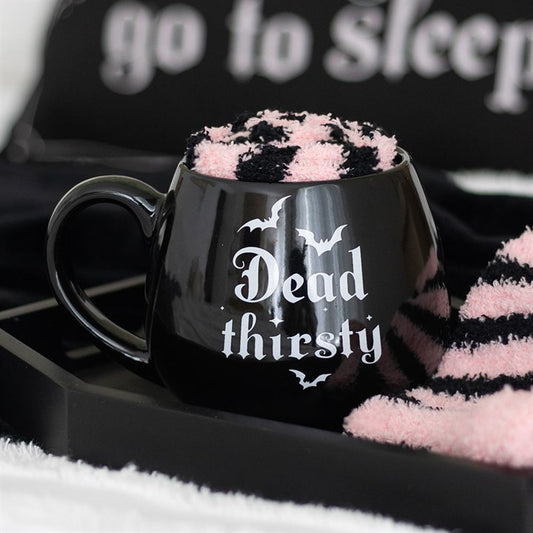 A ceramic mug featuring white Gothic text that reads Dead thirsty and bat illustrations, filled with a set of fluffy pink and black striped socks, sits on a black tray. Part of a sign reading go to sleep is visible in the background.