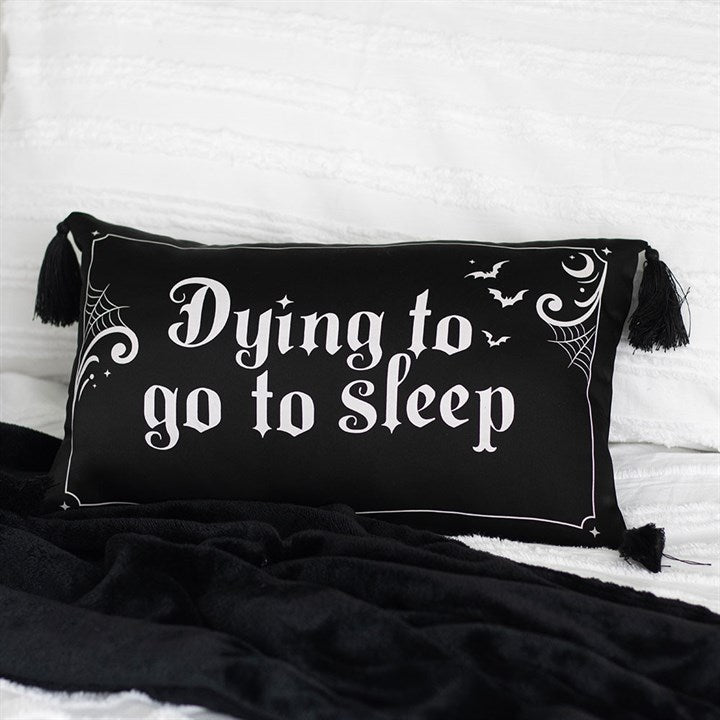 A black rectangular polyester satin cushion with white Gothic lettering reads “Dying to go to sleep.” Spider webs, bats, and stars add gothic comfort, while black tassels on each corner complete this edgy décor piece atop a white bedspread.