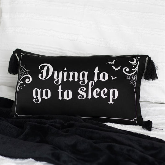 A black rectangular polyester satin cushion with white Gothic lettering reads “Dying to go to sleep.” Spider webs, bats, and stars add gothic comfort, while black tassels on each corner complete this edgy décor piece atop a white bedspread.