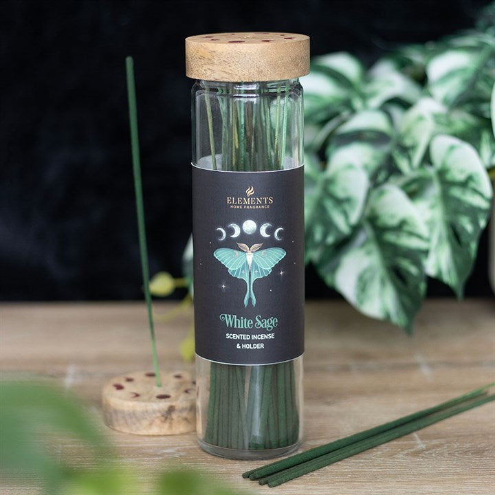 A glass jar with a wooden lid contains green incense sticks labelled “White Sage”, featuring a luna moth design and phases of the moon. A leafy plant and a round incense holder with sticks are in the background.