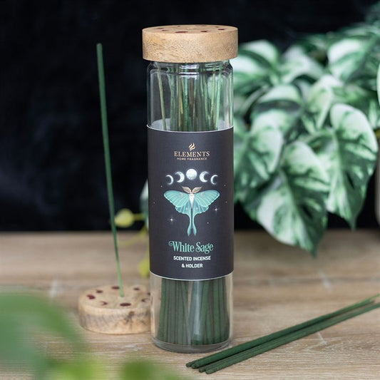 A glass jar with a wooden lid contains green incense sticks labelled “White Sage”, featuring a luna moth design and phases of the moon. A leafy plant and a round incense holder with sticks are in the background.