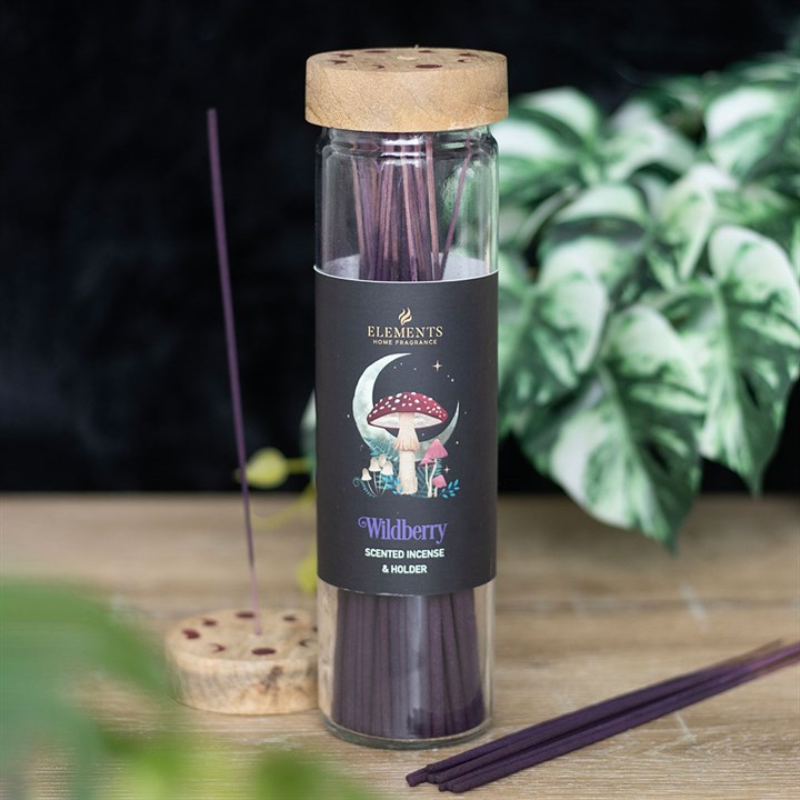 A clear glass jar with a wooden lid holds wild berry fragrance incense sticks. The label features a mushroom, moon, and stars. In the background, a plant with green and white leaves appears blurred.