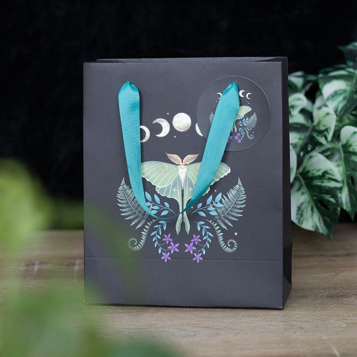 A black gift bag embellished with an illustration of a mint-green luna moth, crescent moons, moon phases, and ferns interspersed with purple flowers. The design adorns both the bag and a circular sticker. Teal ribbon handles enhance its charm as it sits on a wooden surface with green plants in the background.