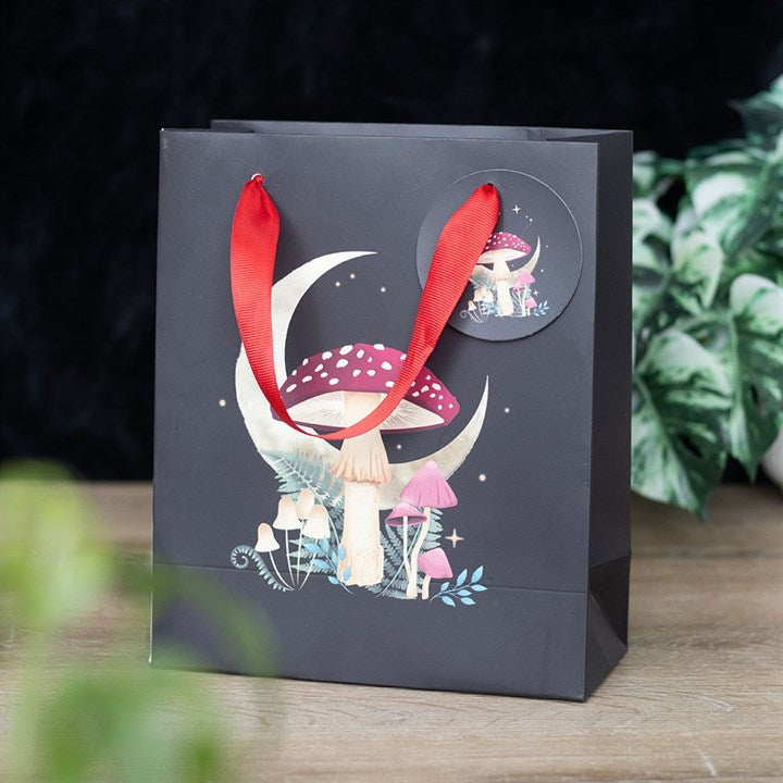 A black gift bag with red handles features a mystical mushroom design beneath a crescent moon. The whimsical illustration showcases red-capped and white mushrooms amidst a dark forest theme. A matching circular tag with the same artwork is attached to the handle, set against lush greenery.
