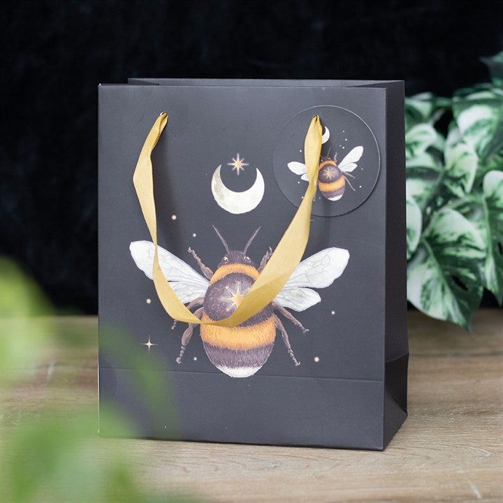 This printed gift bag features a large bee illustration amidst a mystical forest backdrop, with a crescent moon and stars. It has yellow ribbon handles and a matching circular tag showcasing the bee, all set against green foliage.