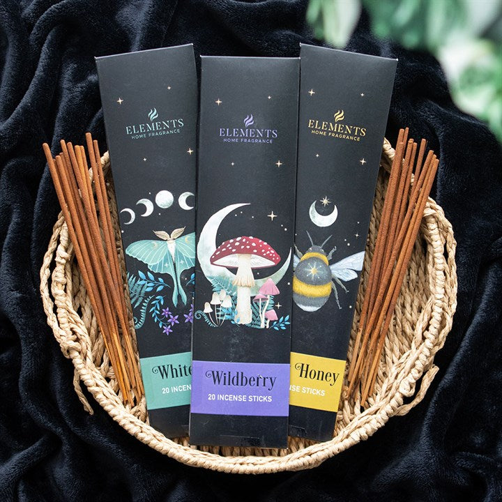 Three boxes of Elements Home Fragrance incense sticks—White Sage, Wildberry, and Honey—stand in a wicker basket filled with brown incense sticks, displayed on black fabric with leafy greenery to make the perfect gift set.