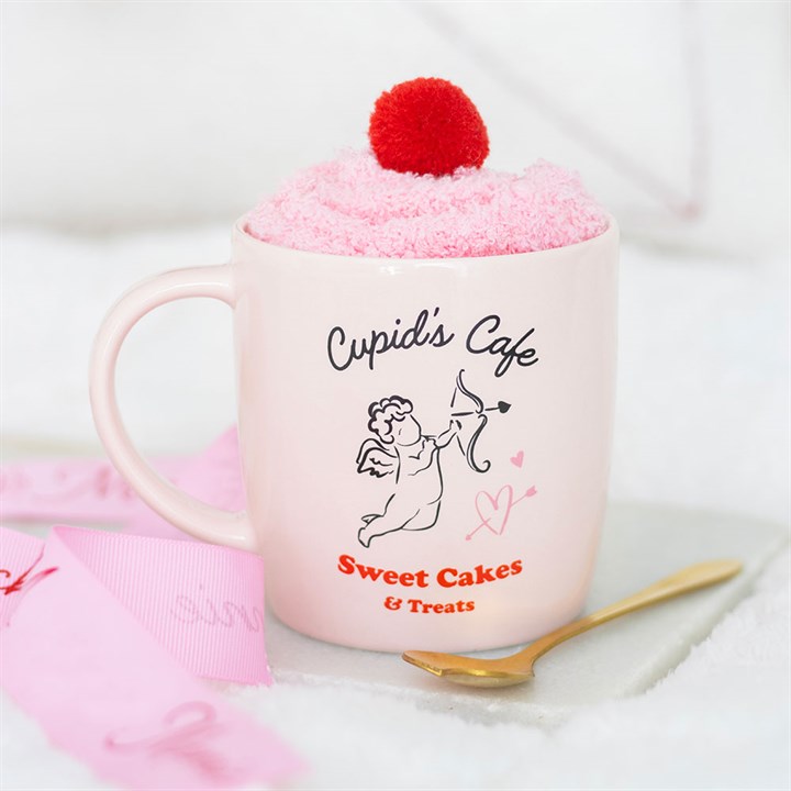 A pale pink Cupids Cafe mug labelled Sweet Cakes & Treats with a cupid illustration holds a fluffy pink dessert topped with a red pom-pom—ideal for a Valentines Day gift set. A gold spoon and pink ribbons lie nearby on a white surface.