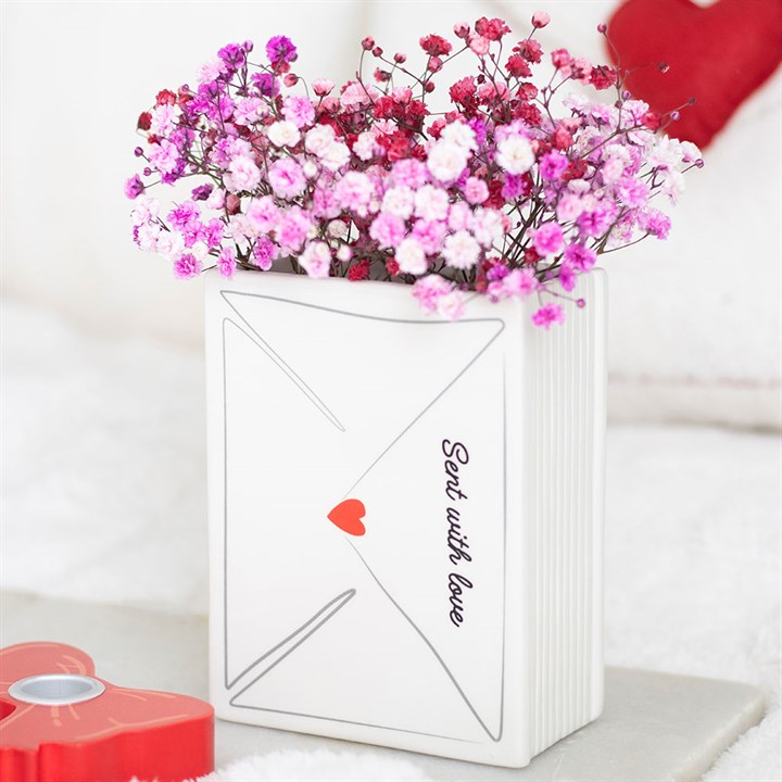 A white ceramic vase shaped like an envelope, marked Sent with love and decorated with a red heart, contains pink and purple babys breath flowers—a lovely Valentine’s Day decoration or thoughtful gift. A red heart shape is in front.