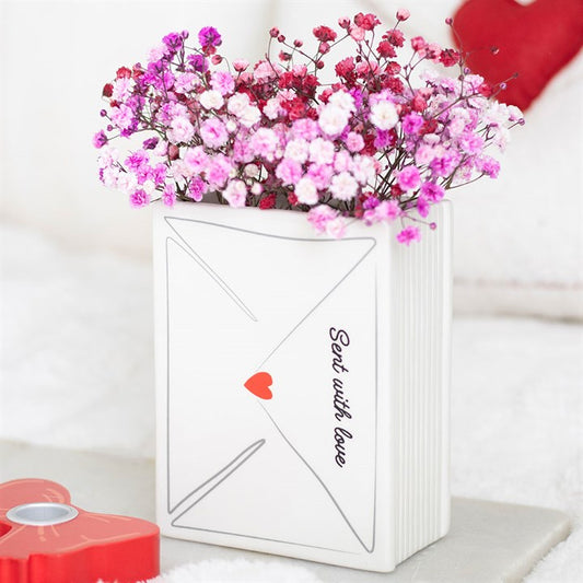 A white ceramic vase shaped like an envelope, marked Sent with love and decorated with a red heart, contains pink and purple babys breath flowers—a lovely Valentine’s Day decoration or thoughtful gift. A red heart shape is in front.