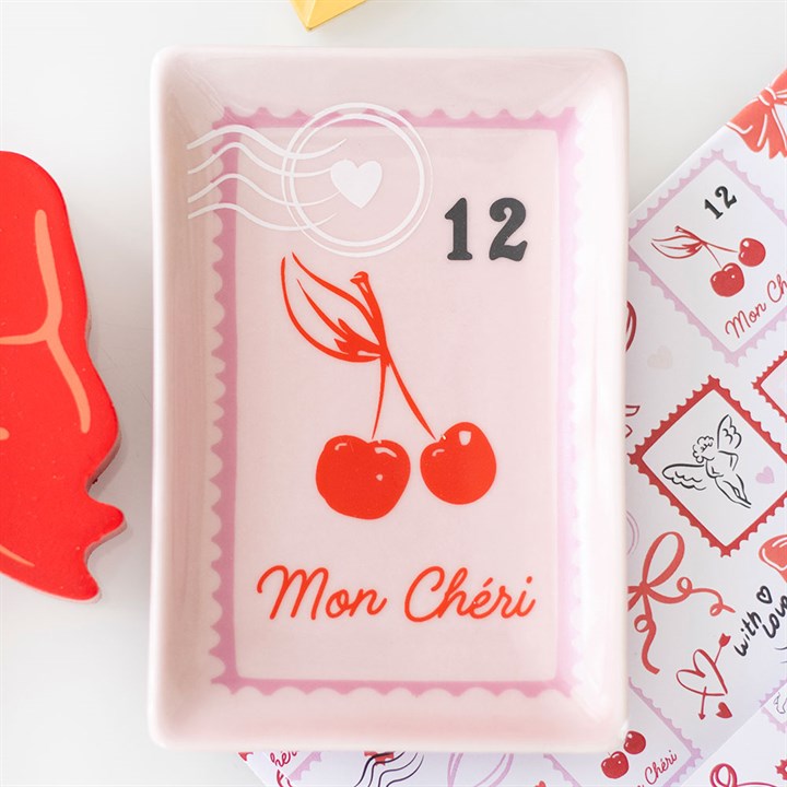 A rectangular pink trinket tray displays two French cherries with leaves, the number 12, a white postage stamp motif with a heart, and Mon Chéri written in red cursive at the bottom.