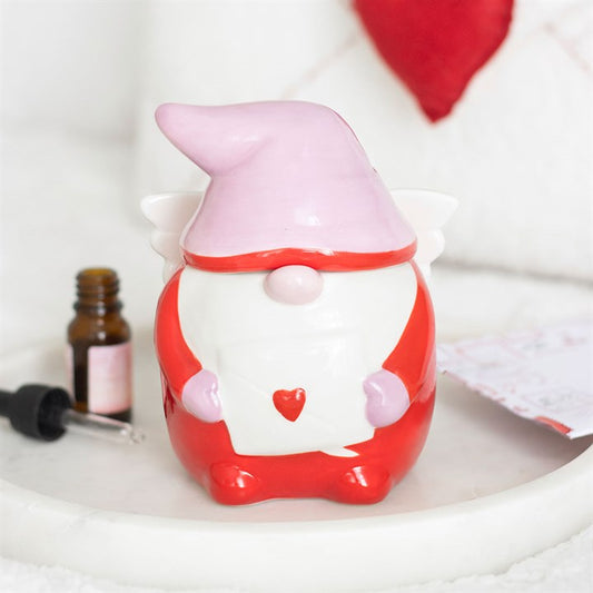 A ceramic gnome ornament with a pink hat, red suit, and white beard holds a white envelope featuring a red heart. It stands next to fragrance oil on a tray, with a red heart cushion in the background—ideal for Valentine’s Day.