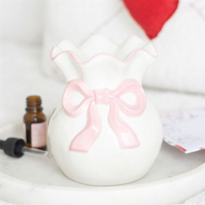 A white ceramic vase with a ruffled rim and pink ribbon motif sits in a coquette style on a marble tray. A small dropper bottle is nearby, with a note and red cushion in the background—ideal Valentine’s Day décor.